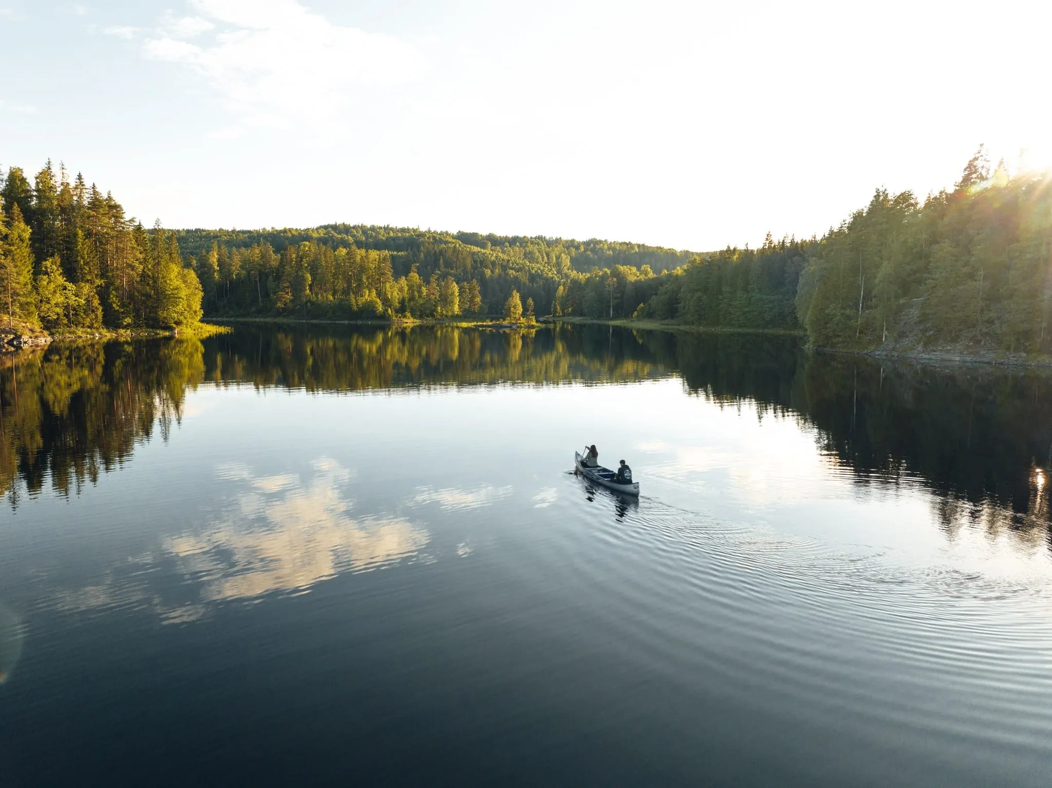 A canoe with two paddlers on a still forest lake, clouds reflected in the water