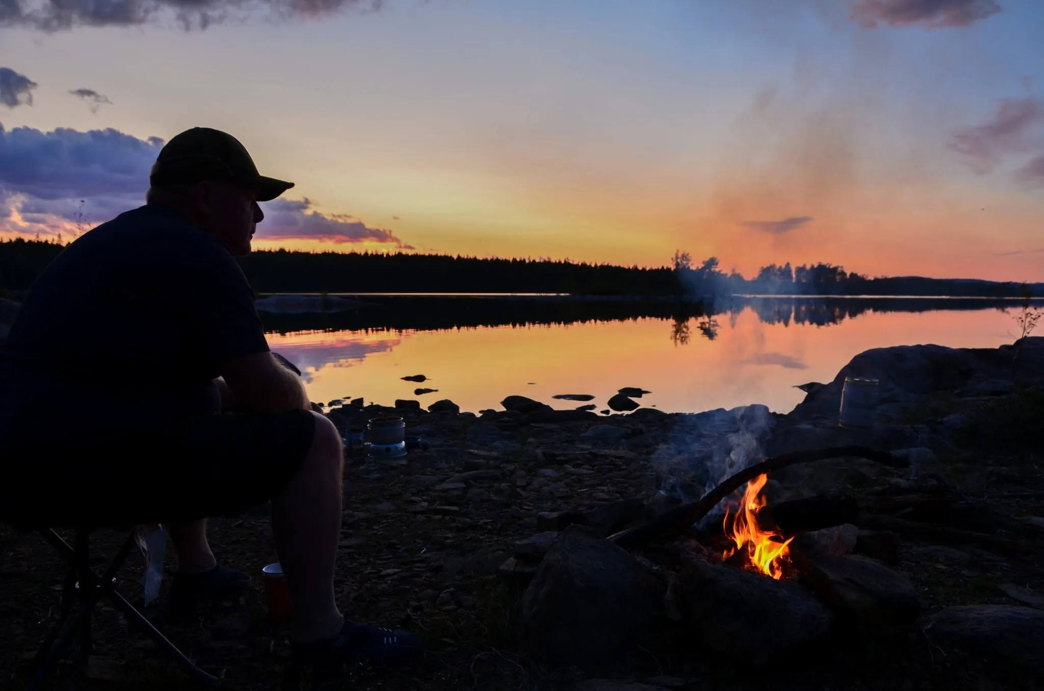 A paddler sitting by a campfire on a rocky lakeshore at dusk