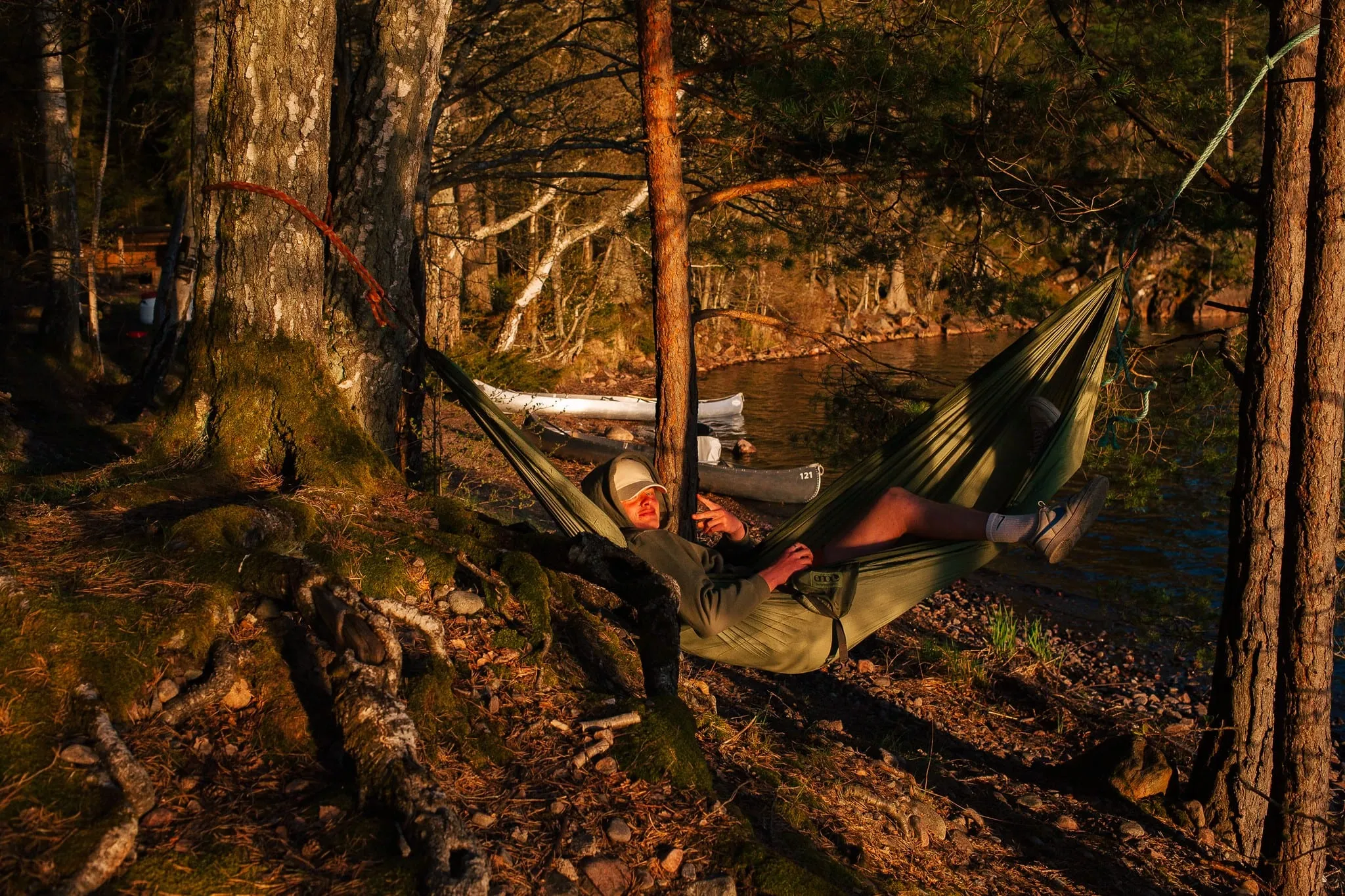A camper resting in a hammock between pine trees by a lake in the evening sun